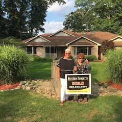 Jan Jacobs and an older woman posing next to a sold sign on a green lawn and a brick house